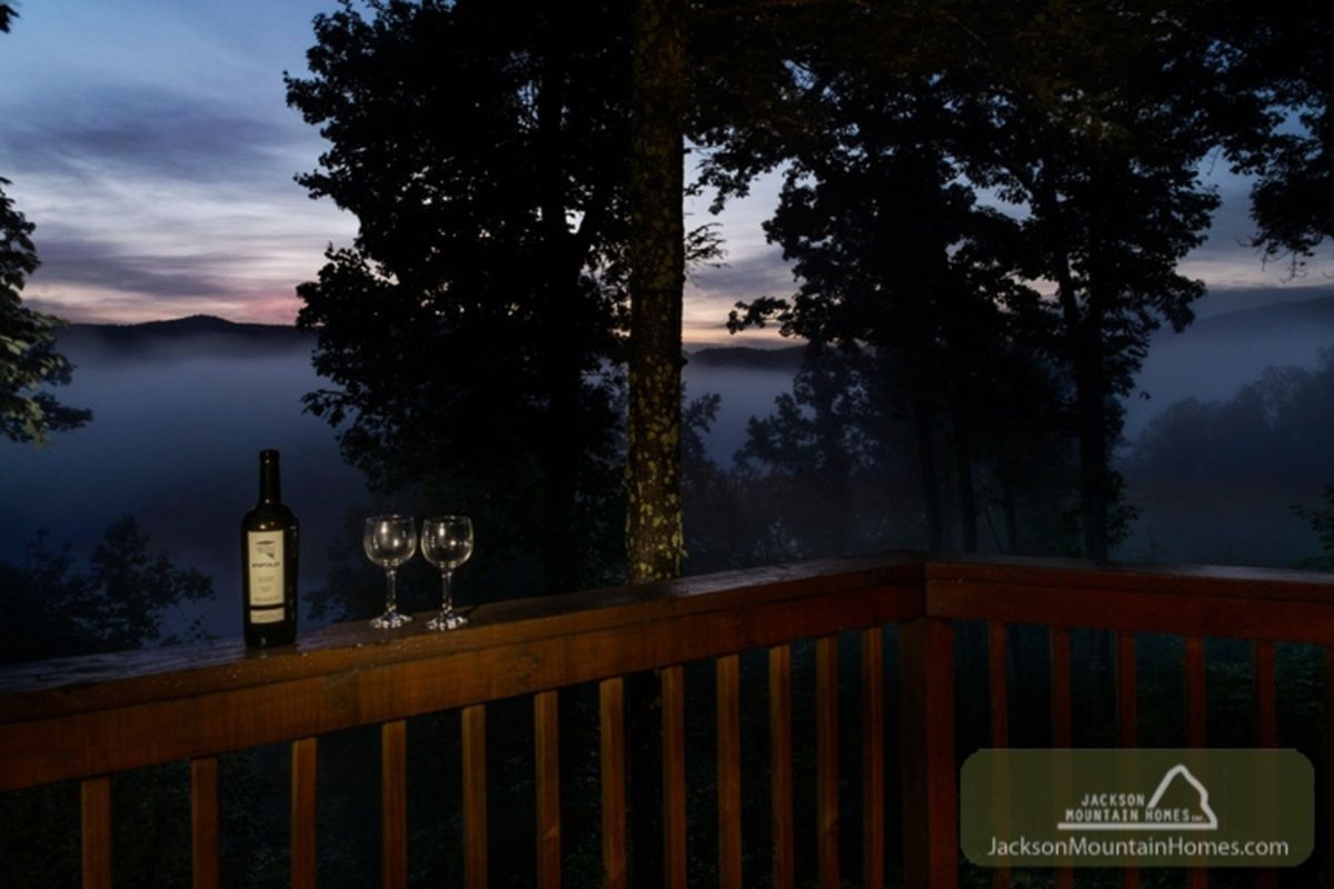Fog rises amid the blue Smoky Mountains just after sunset with the sky still tinged purple in the distance. On the cabin's patio, a bottle of red wine and two glasses await, adding to the air of romance.